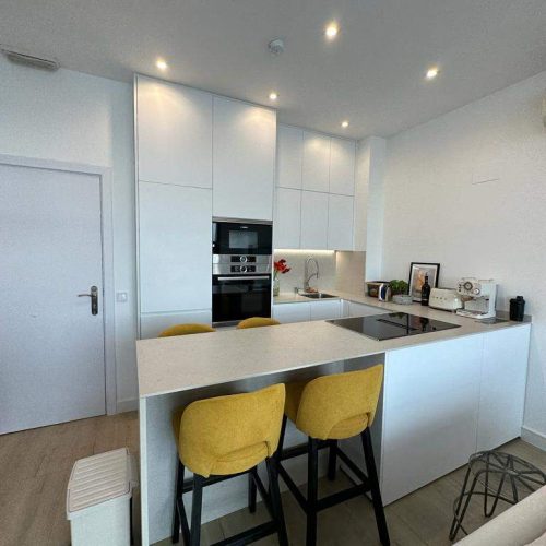 Cocina blanca con isla central y taburetes amarillos diseñada en reforma integral en Madrid